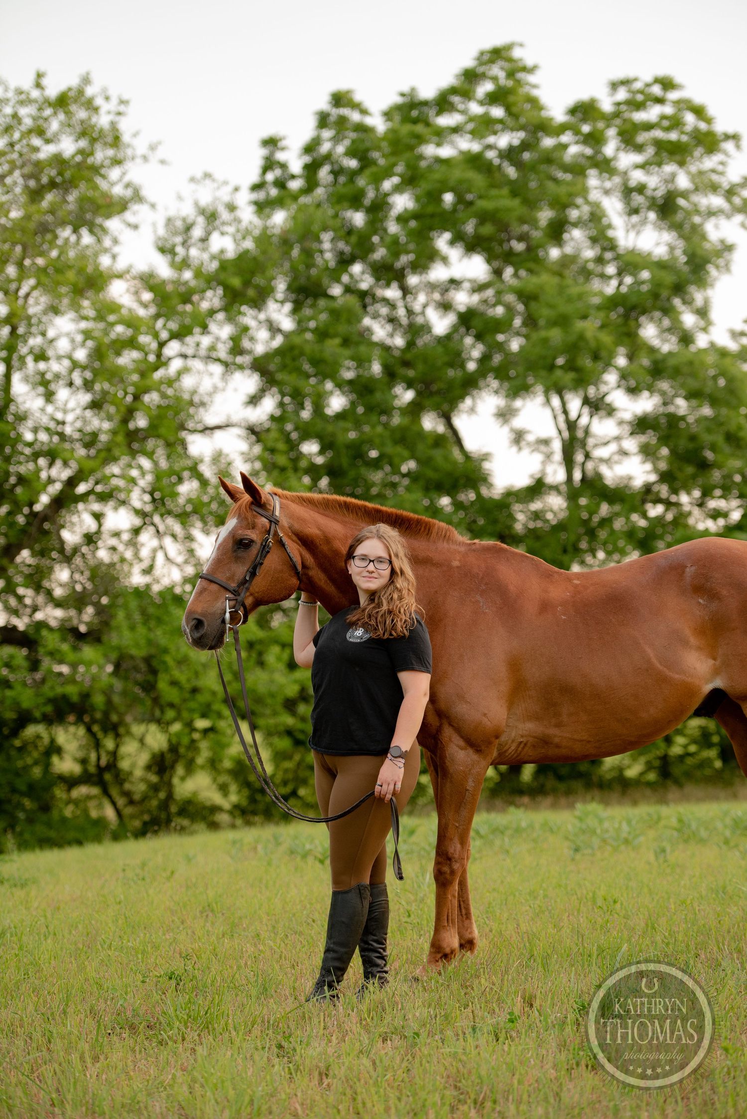 A beautiful chestnut horse poses with its handler in riding boots against a verdant summer landscape.