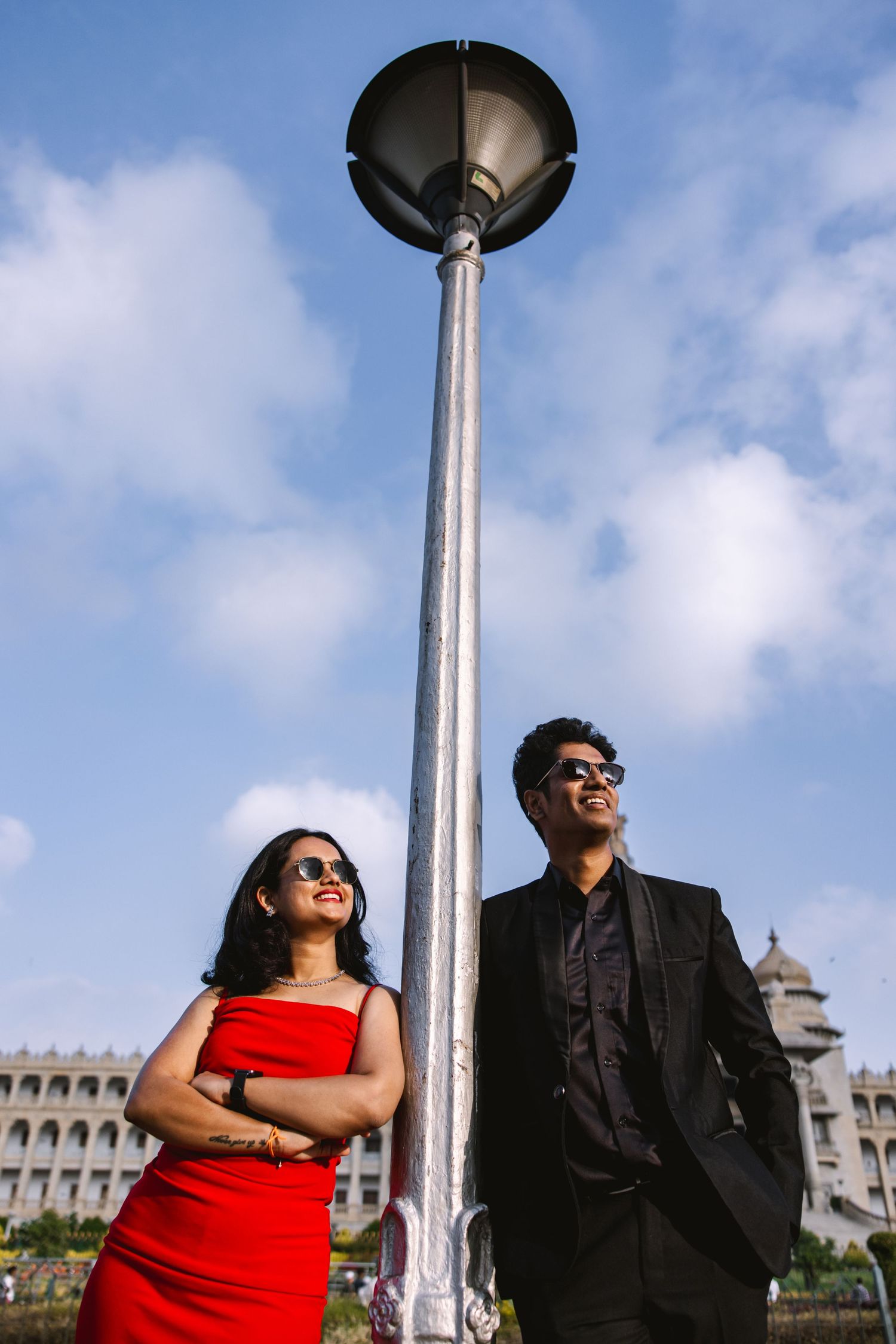 Stylish couple posing against urban backdrop with lamp posts and blue sky.