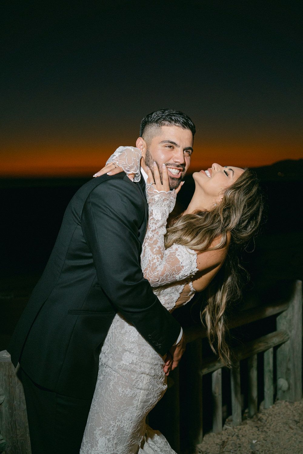 A playful wedding couple shares a dramatic dip pose against a sunset sky.