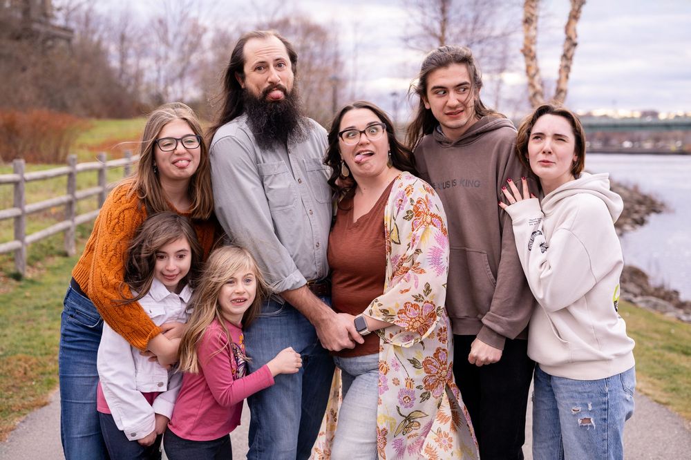 A group of people standing together outdoors on a cloudy day near a waterfront with a fence and trees in the background.