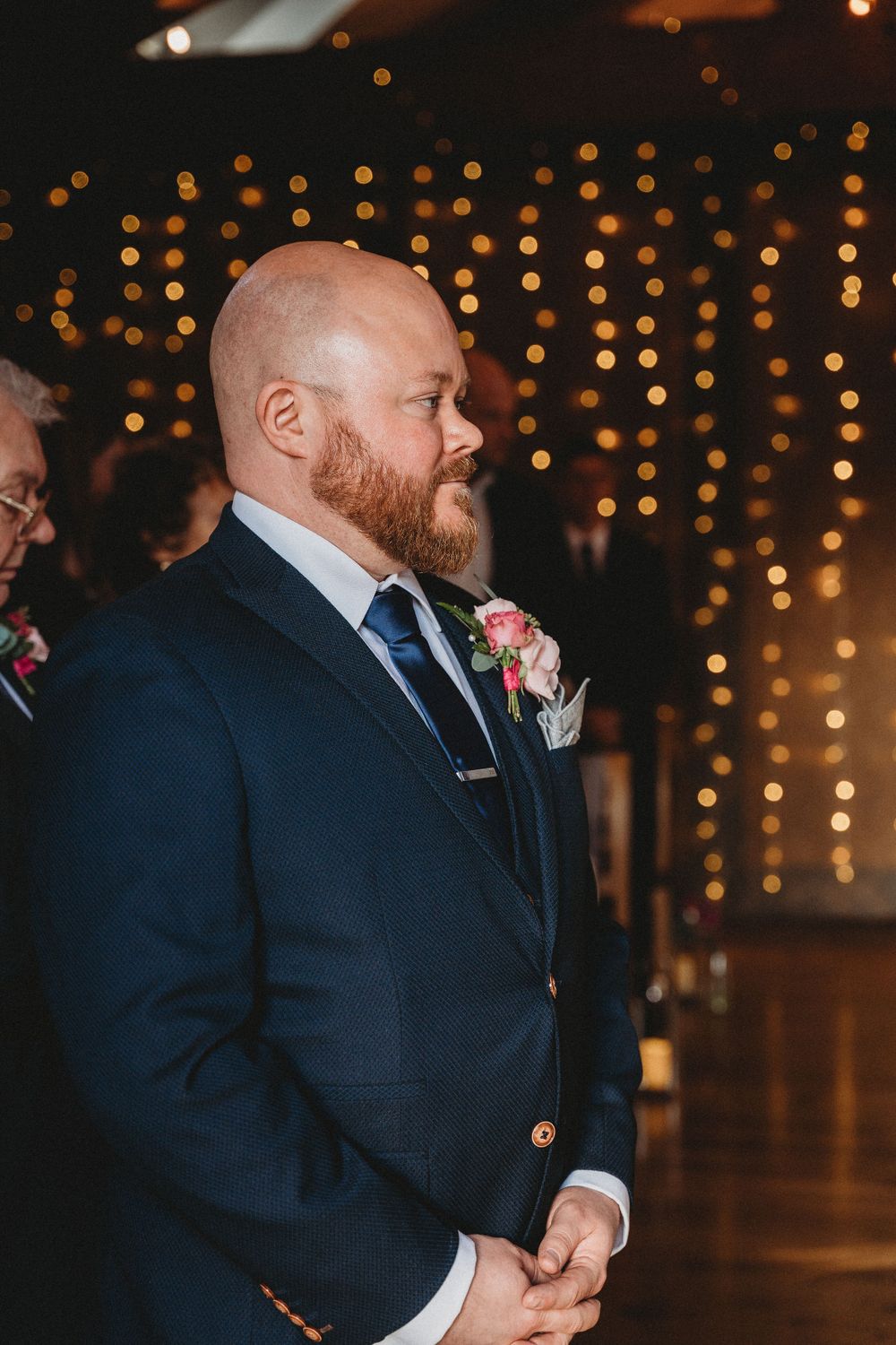 Groomsmen in navy suits stand during wedding ceremony under warm lighting.
