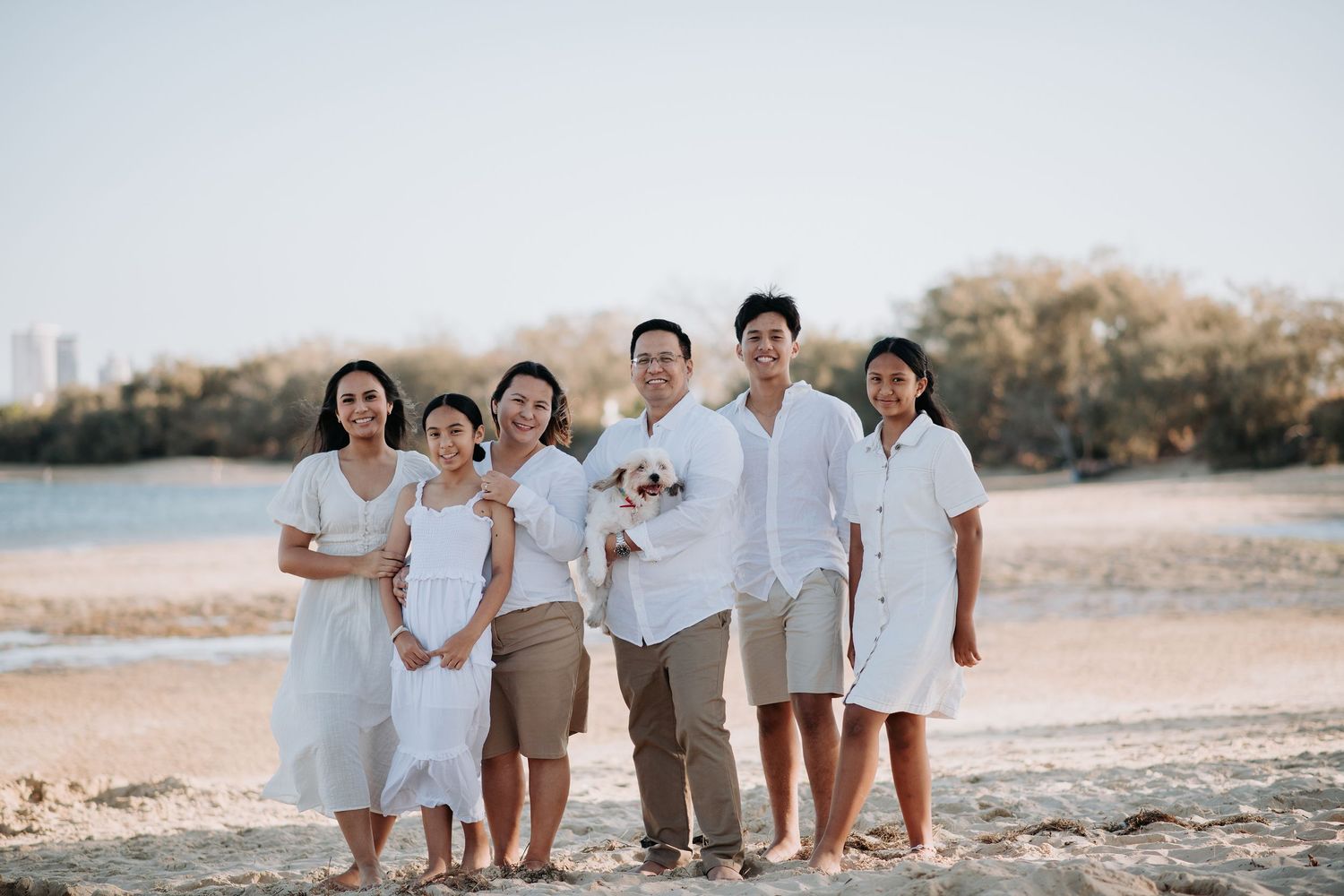 A beachside family portrait with coordinated white outfits against a waterfront backdrop.