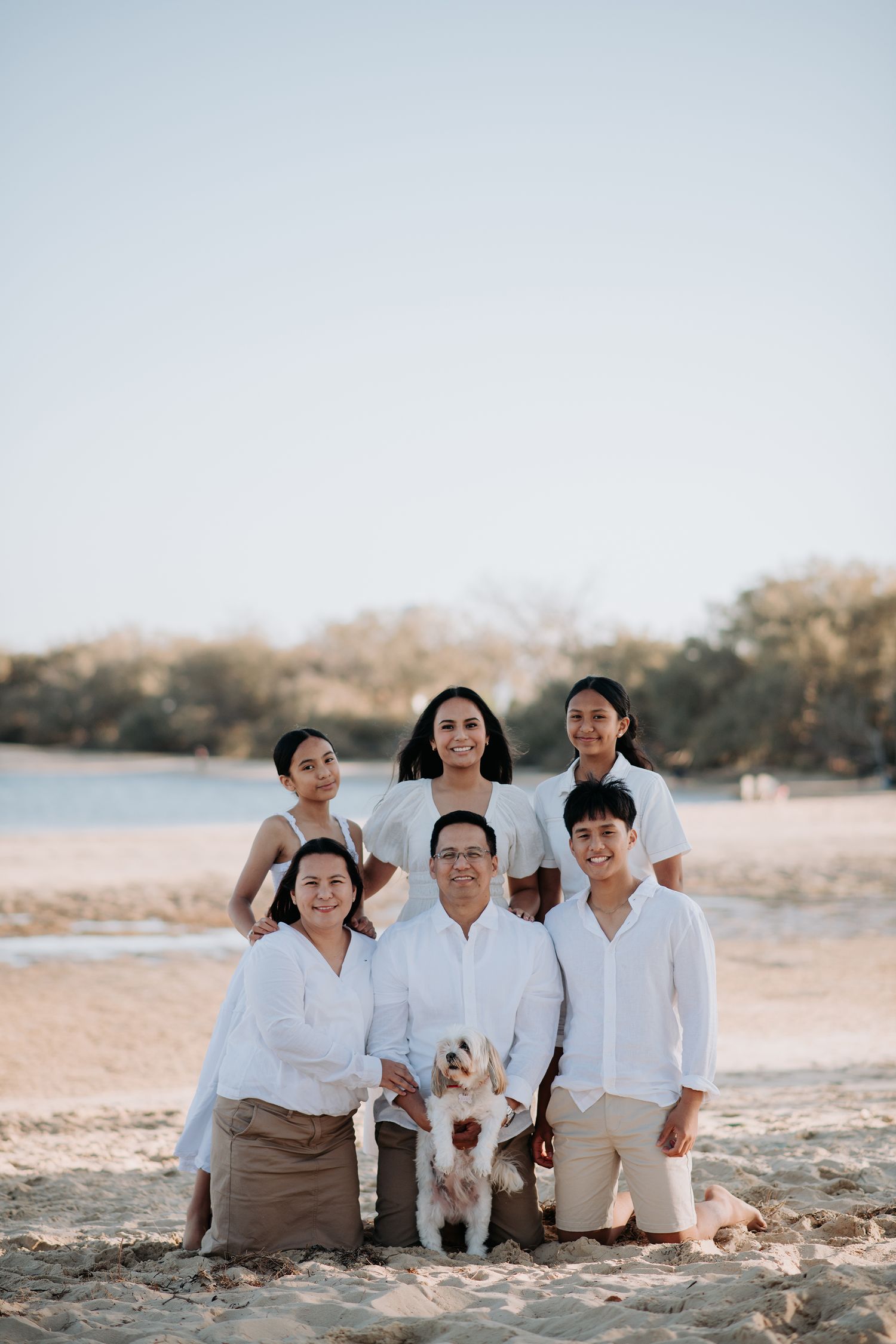 A group portrait at a beach with a small white dog, everyone dressed in light-colored clothing.