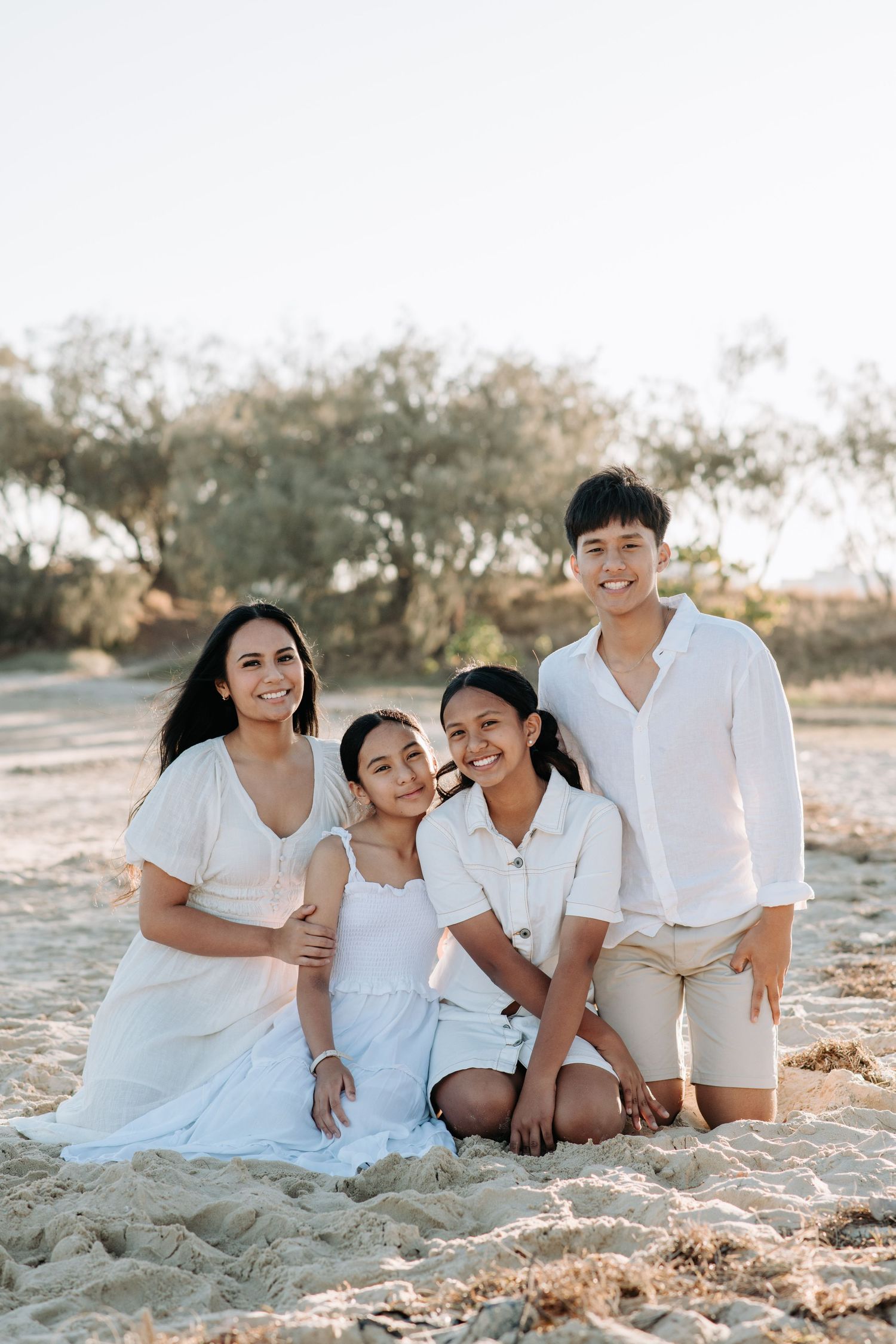 Family of four wearing coordinated white outfits sitting together on sandy beach.