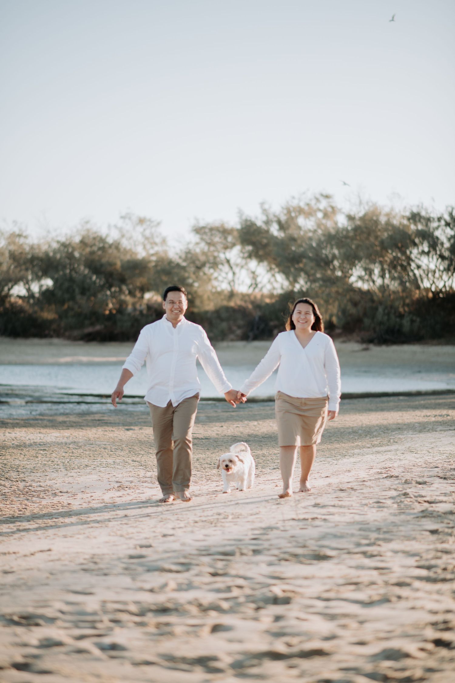 A couple walks their small white dog along the beach path during golden hour.