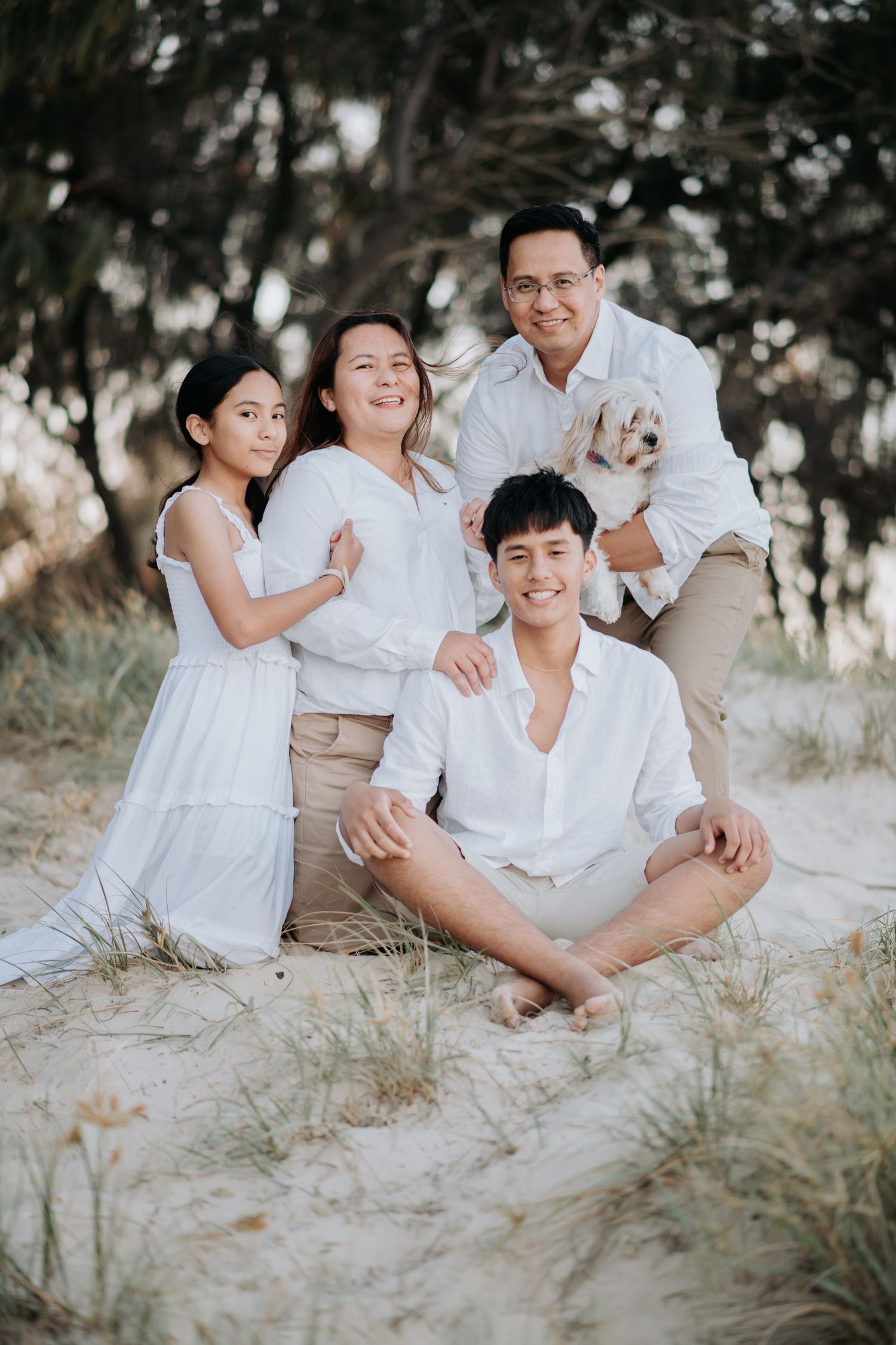 Family group in white clothing sitting together on sandy beach with trees behind them.