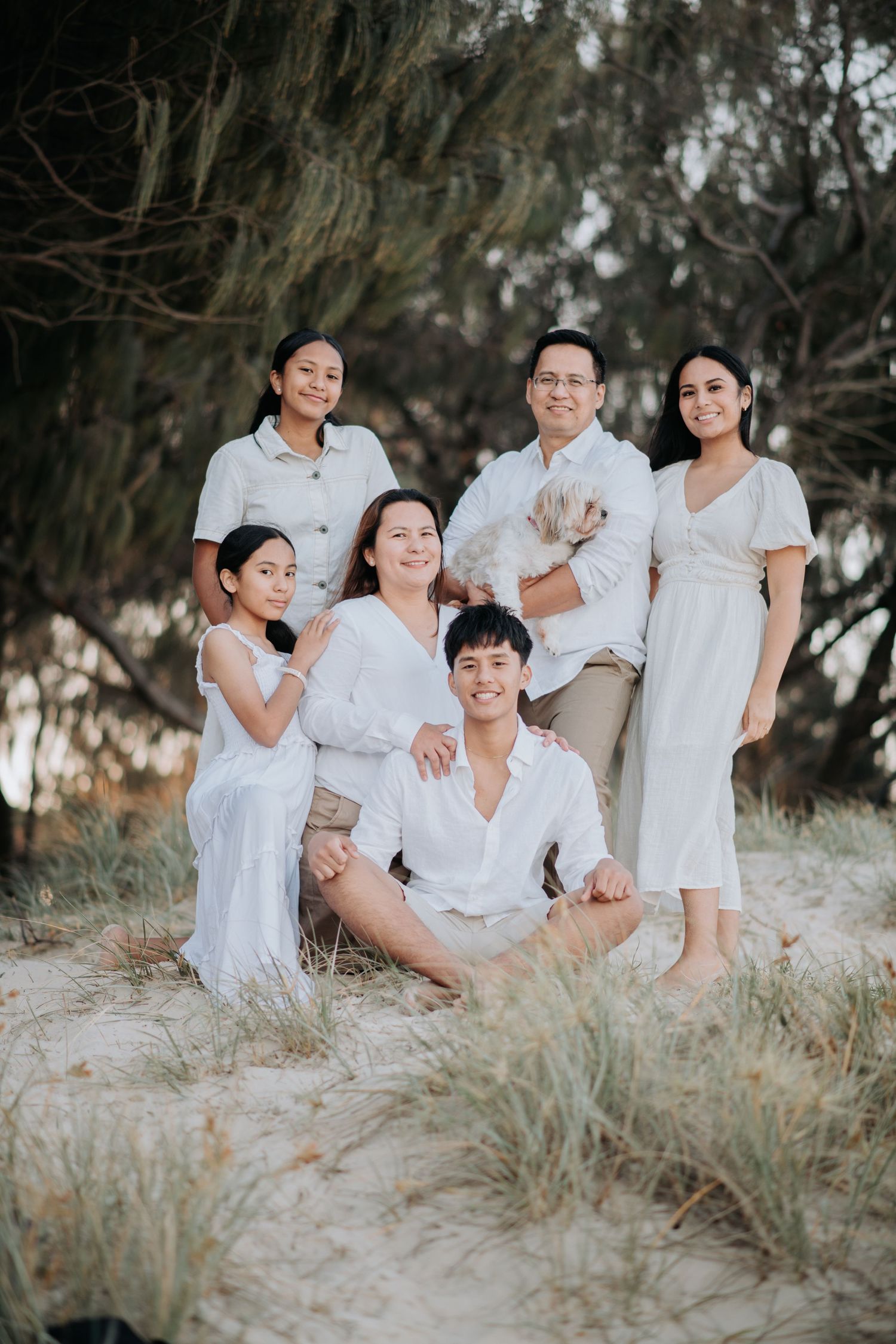 Family of six wearing coordinated white outfits posing together on sandy beach with trees in background.