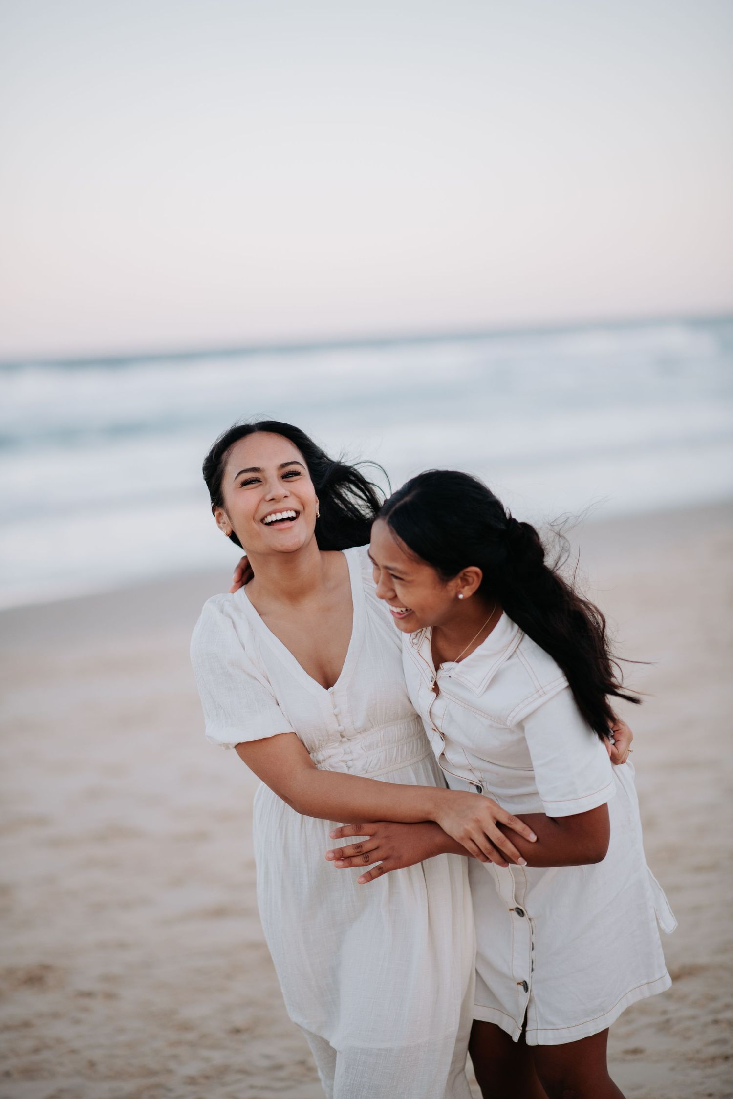 Two people in white outfits laughing and hugging on sandy beach during sunset.