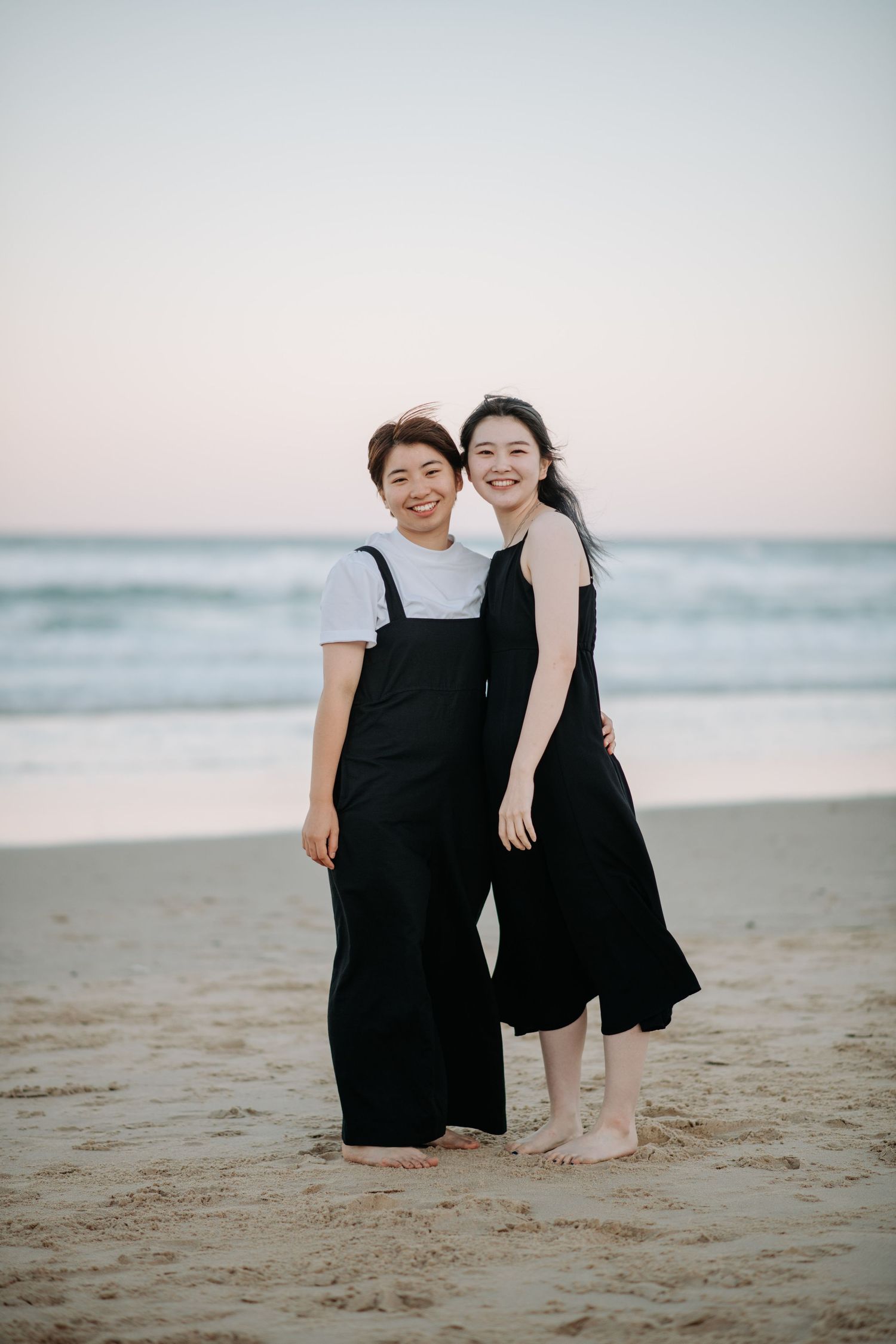 Two people in black dresses share a joyful moment on the beach at sunset.
