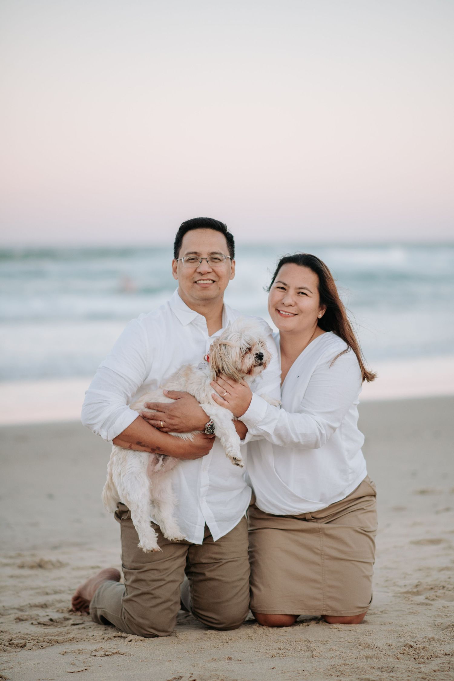 A couple in white shirts holds their small white dog while posing on the beach.