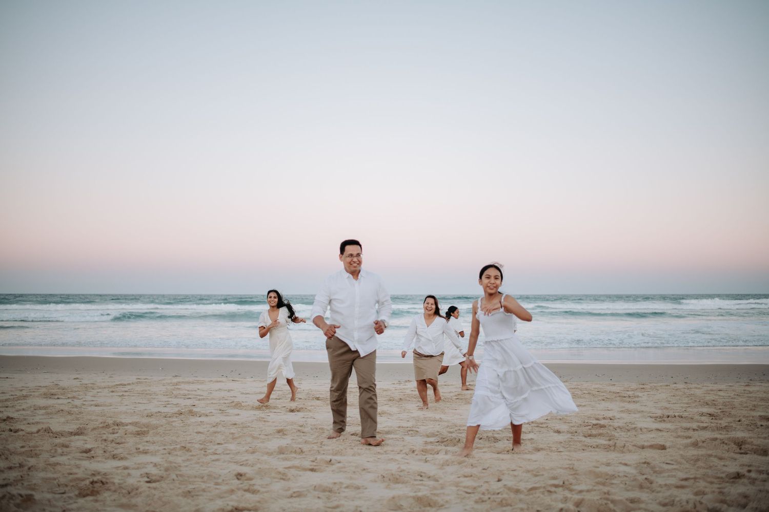 A group runs joyfully along the beach at sunset wearing white outfits.