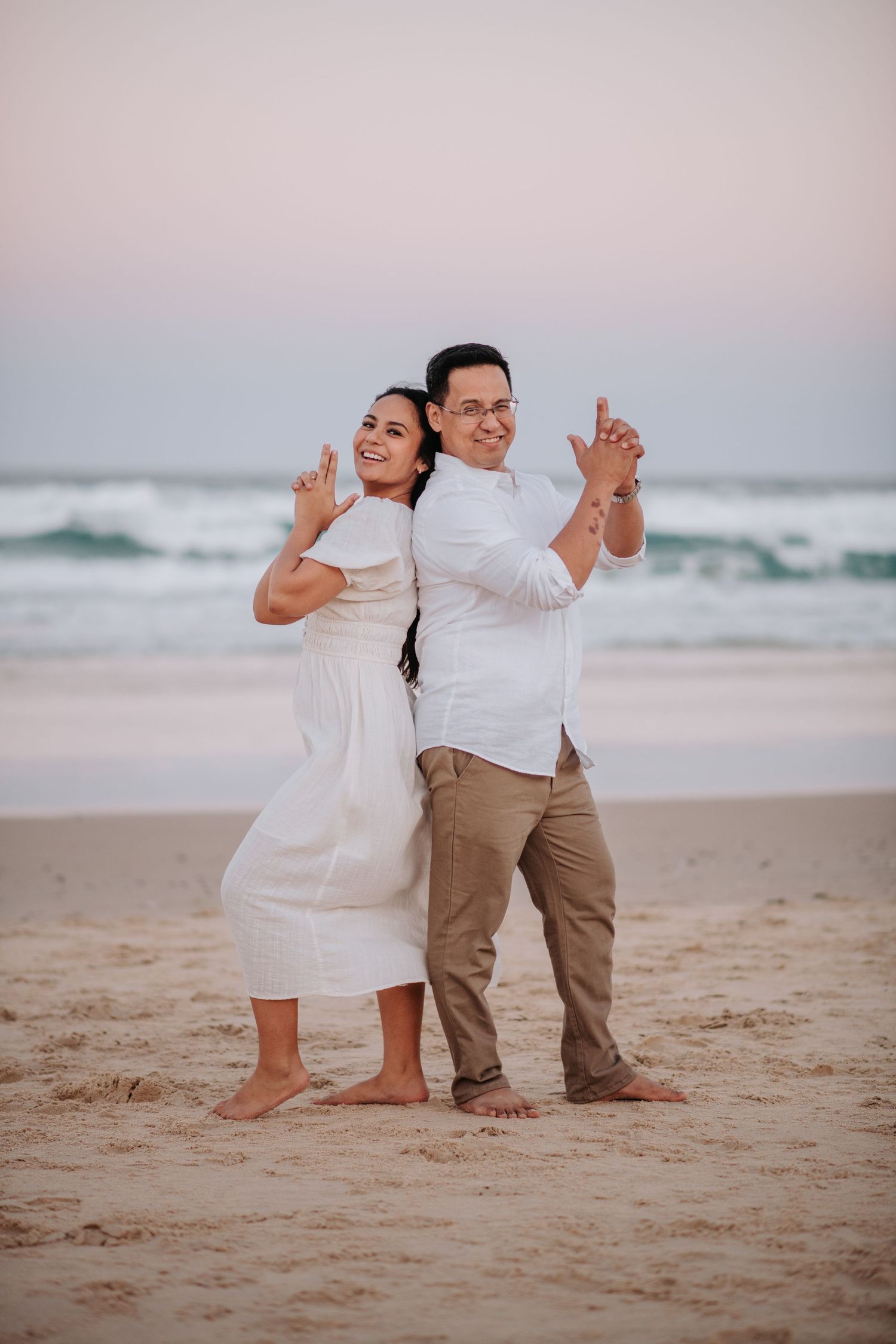 A playful couple poses back-to-back on the beach at sunset in coordinated outfits.