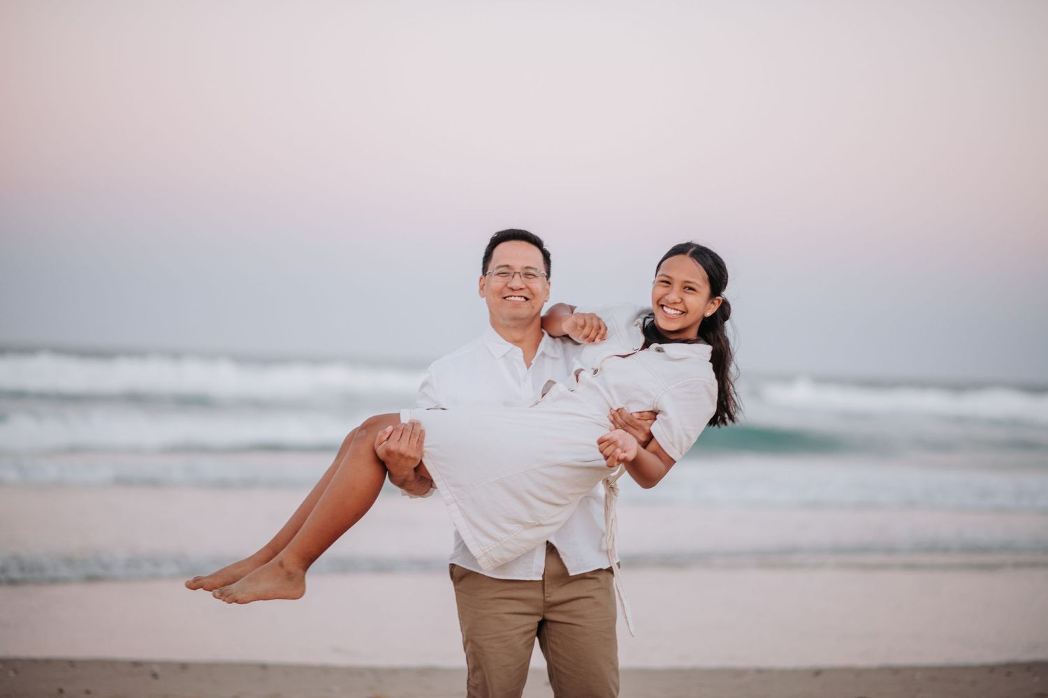 Romantic couple in white clothing embracing on beach at sunset with ocean waves in background.