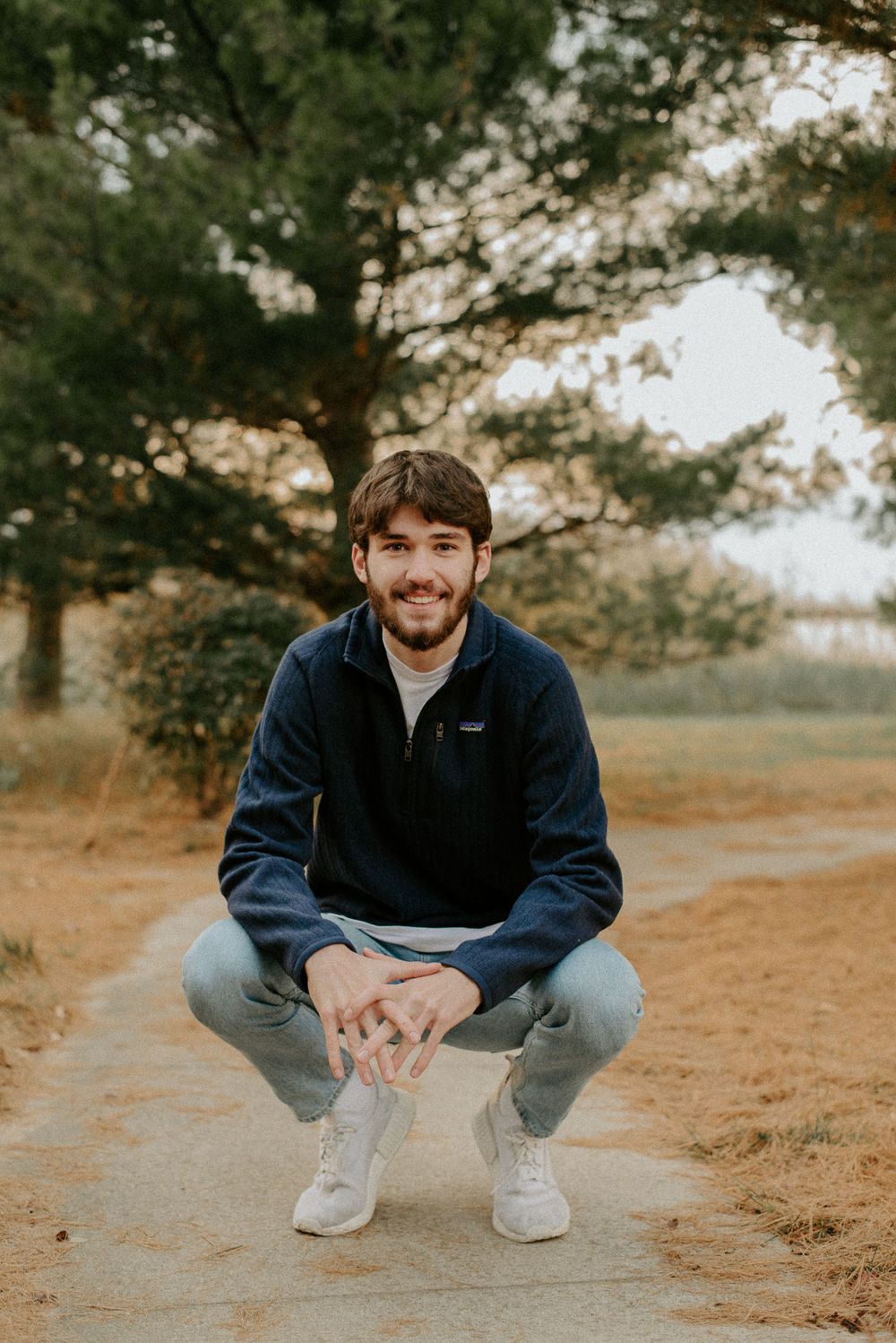 Person in navy blue fleece quarter-zip and jeans crouches on dirt path near pine trees.