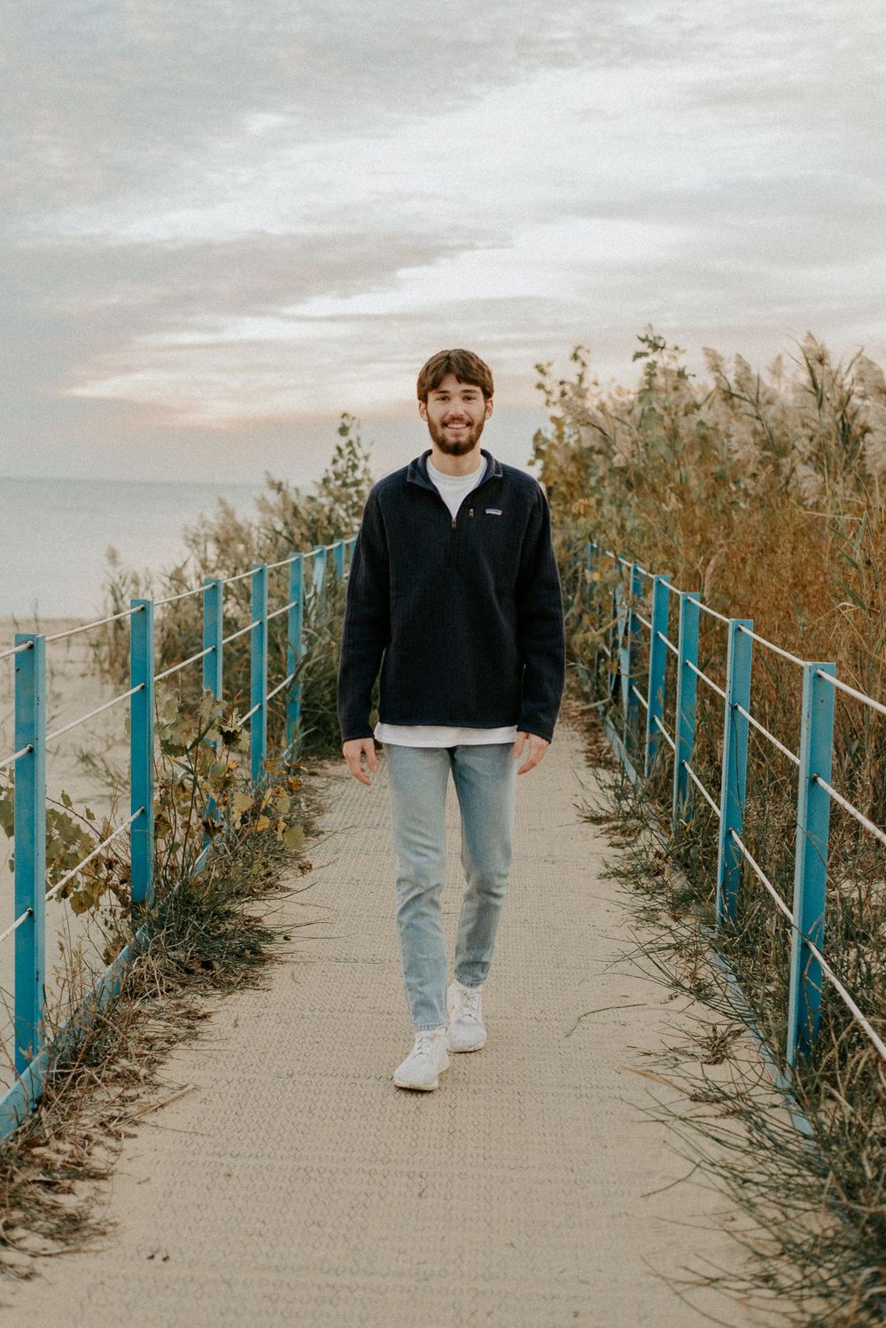 A person walks along a coastal path with blue railings and tall grass at dusk.