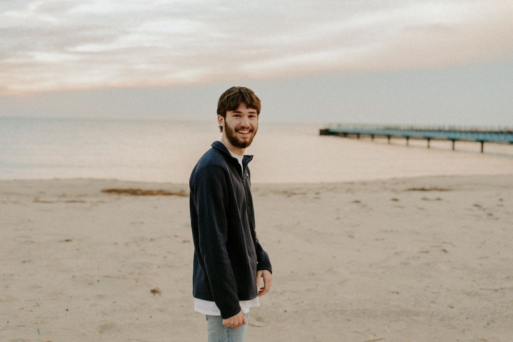 A person in a navy jacket and jeans stands on an empty beach at dusk.
