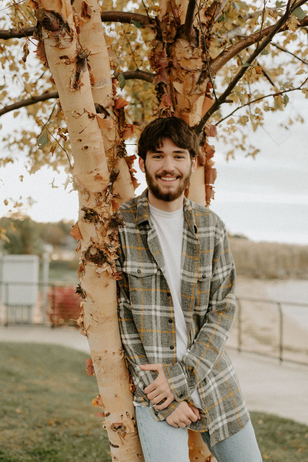 Person in plaid shirt smiles while standing next to birch tree trunk outdoors.