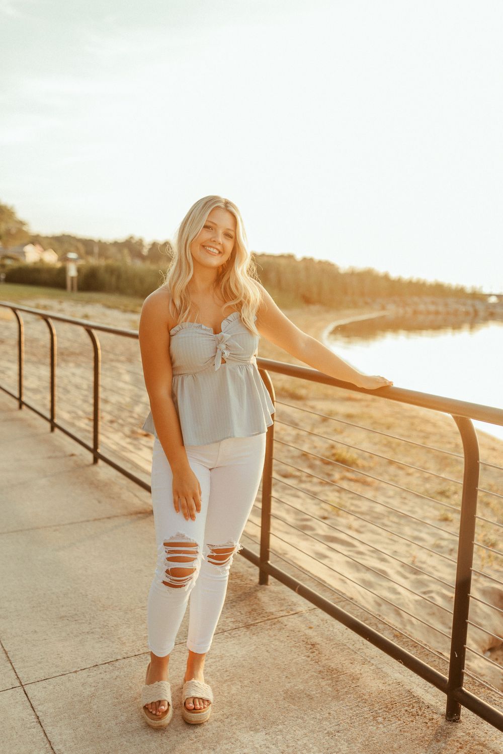 A person in a light blue top and distressed white jeans poses next to a railing at sunset on a beach path.
