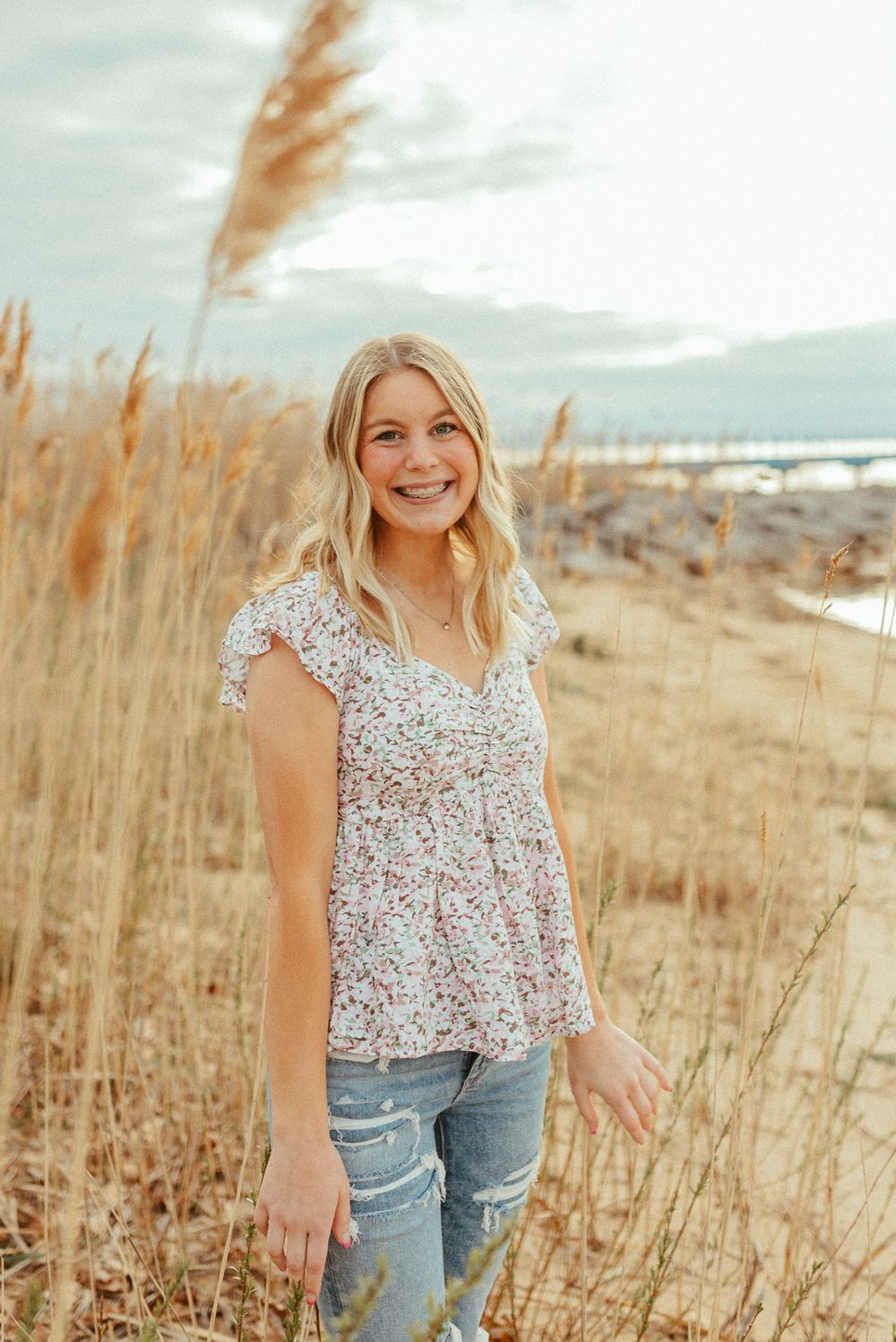 Woman in floral top and distressed jeans poses among tall beach grass at sunset.