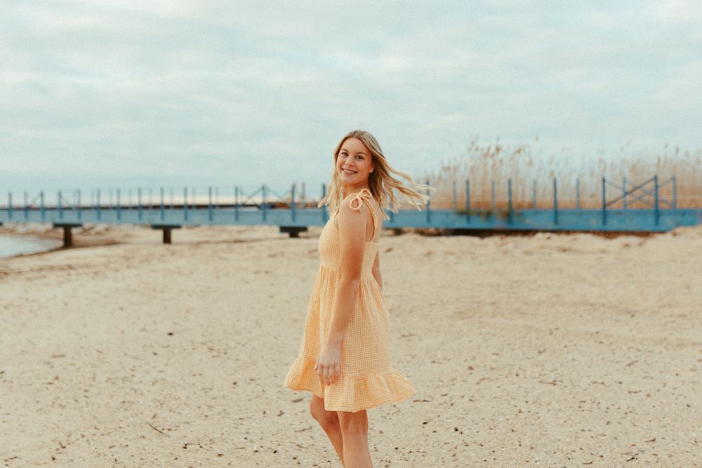 Woman in flowy peach dress twirling on beach with pier in background.
