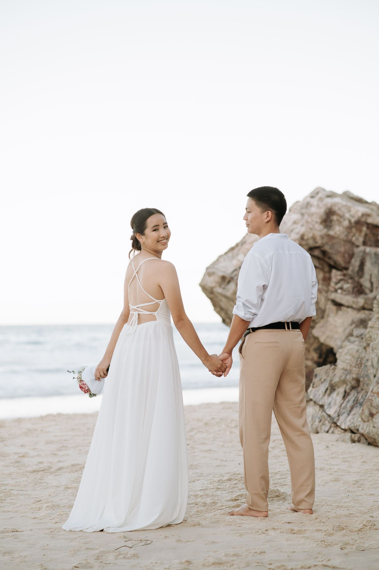 A couple holds hands on the beach with rocky cliffs in the background during sunset.