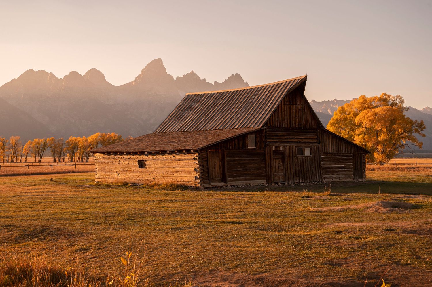 Historic wooden barn sits beneath majestic mountain peaks during golden hour in Grand Teton National Park.