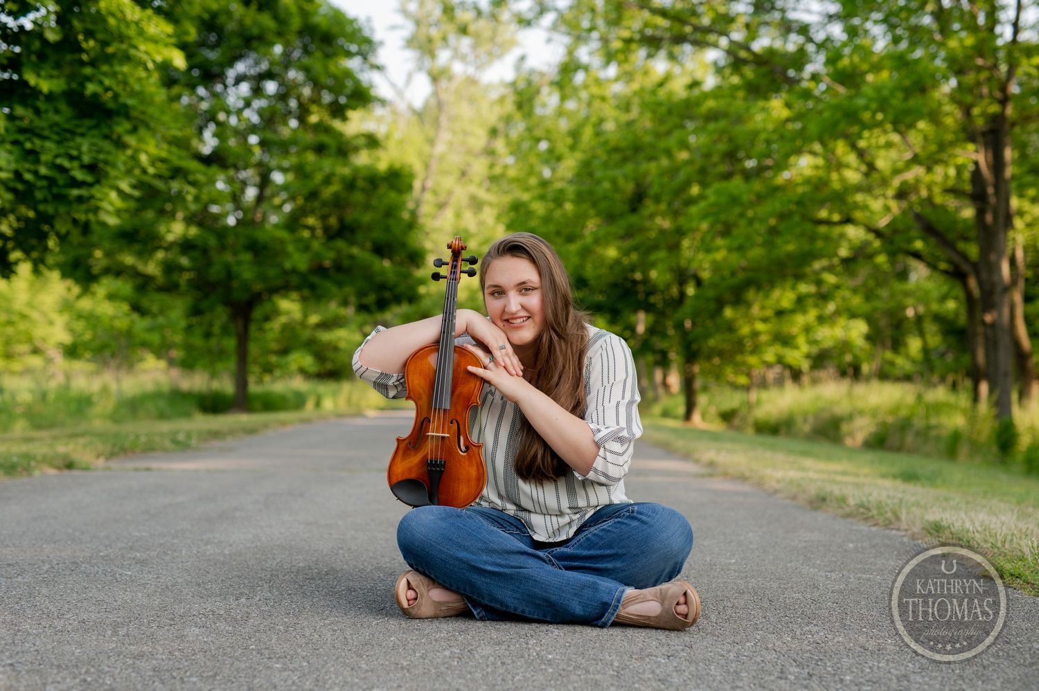 Musician sits cross-legged on path holding violin with trees in background.