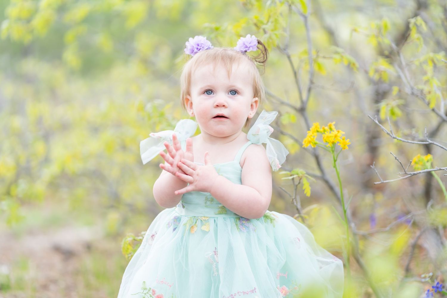 Little girl in mint green dress poses among yellow spring blossoms.