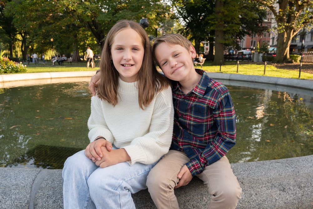 Two people in cozy fall outfits sit together on a stone ledge near a pond in a park setting.
