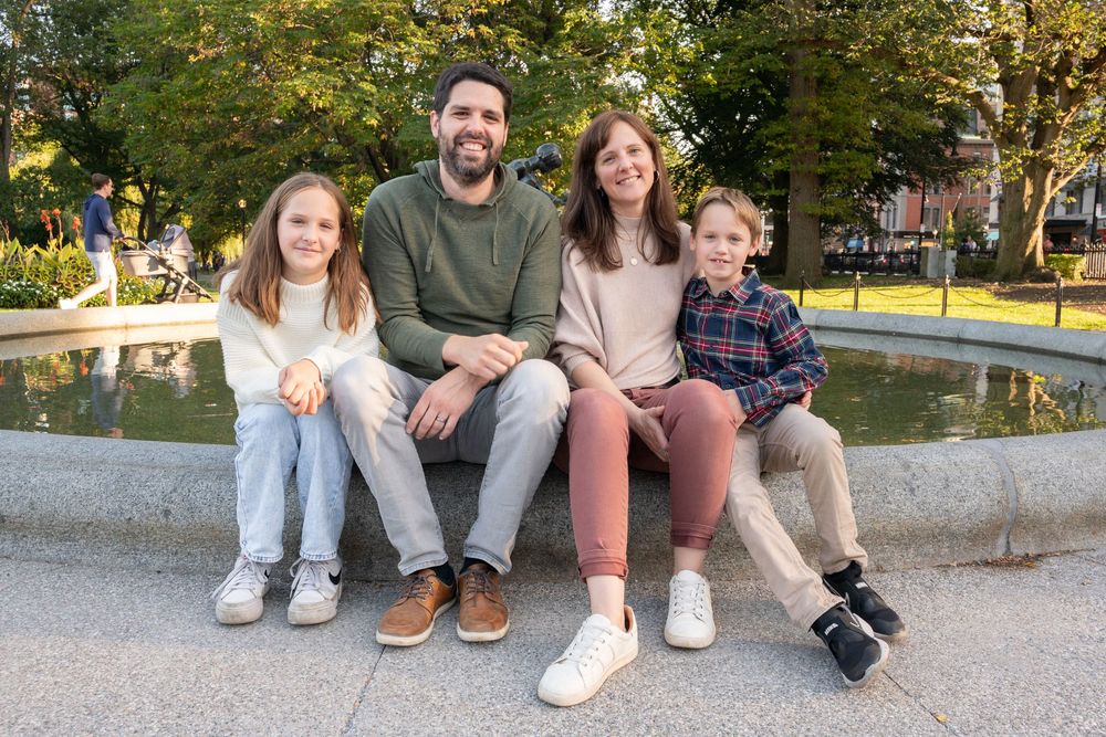 A group sits together on a stone ledge near a fountain in a park during autumn.