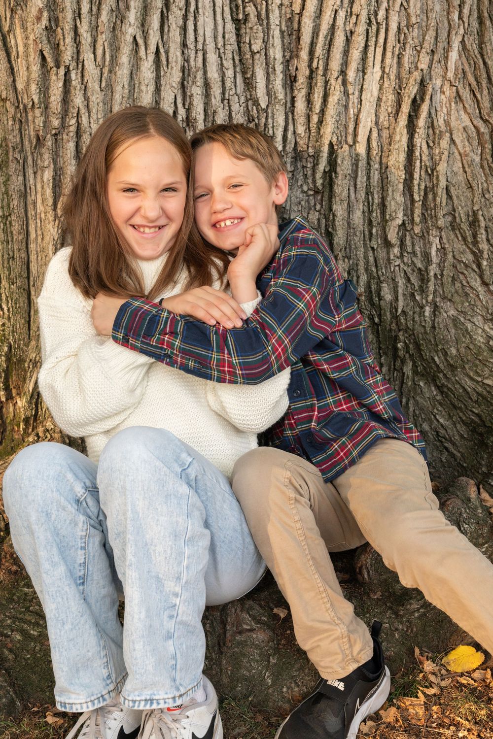 Two children sit together and laugh while leaning against a large tree trunk on an autumn day.