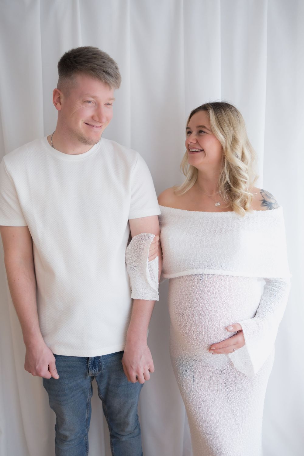 Smiling couple in white outfits, pregnant woman in off-shoulder dress holding belly, man in white tee and jeans, studio backdrop.