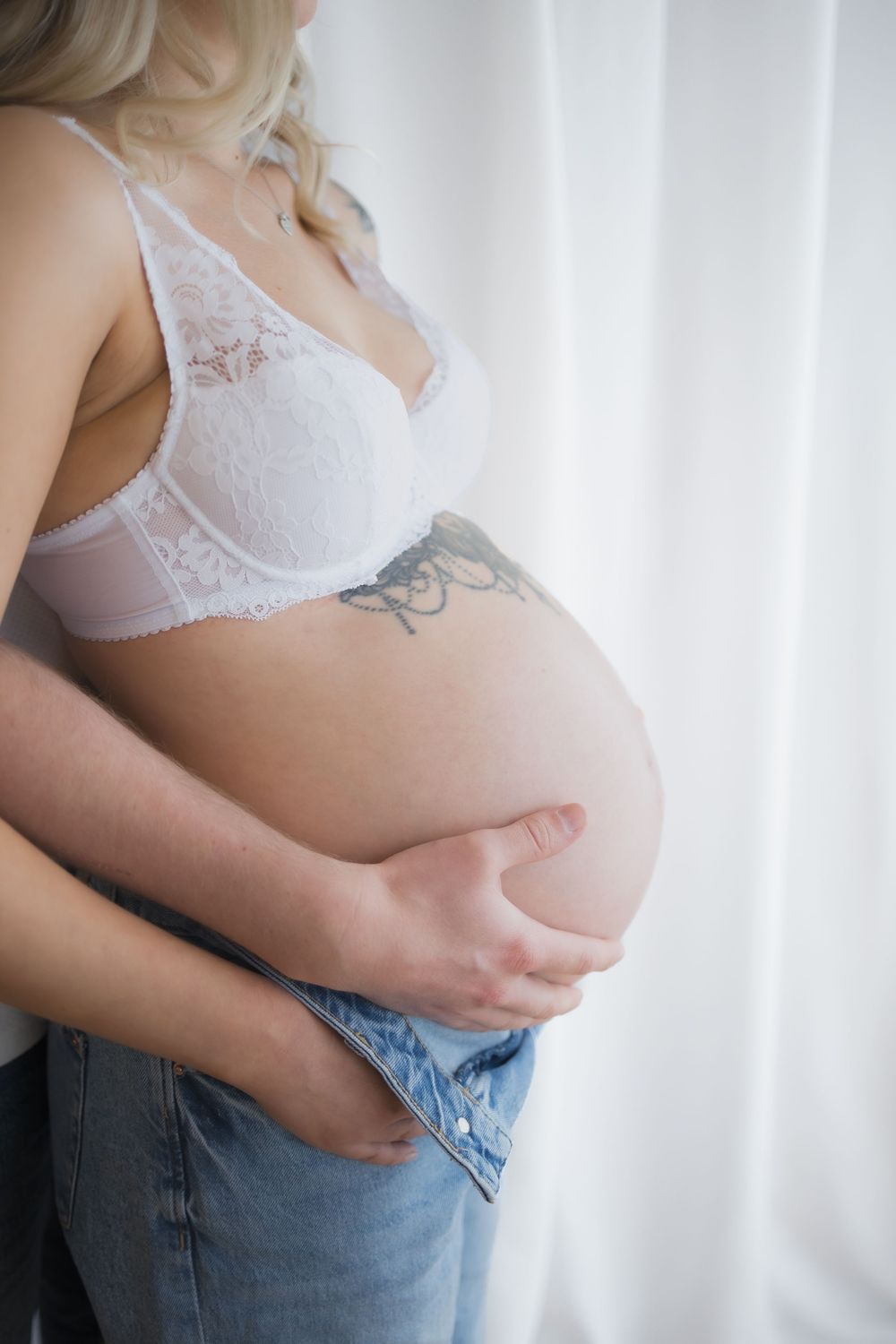 Pregnant woman in white lace bra and jeans cradling her baby bump, showing a floral tattoo on her side.
