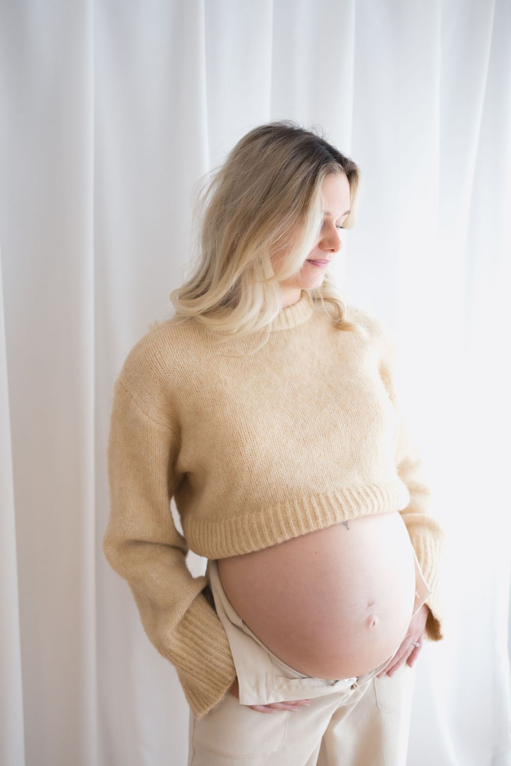 Pregnant woman in beige cropped sweater cradling her bare baby bump against a white curtain backdrop.