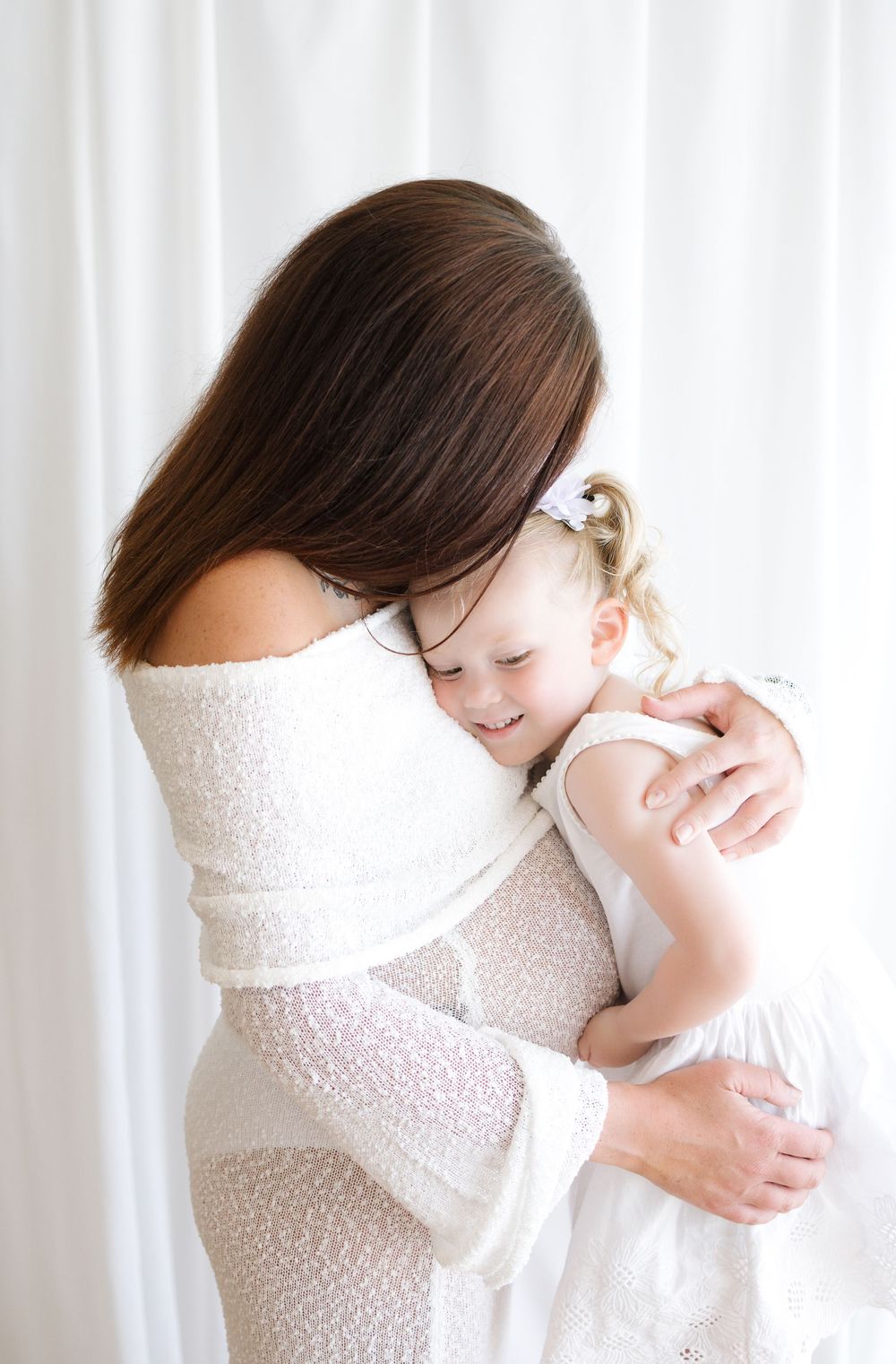 A mother in a white lace dress tenderly embraces her smiling young daughter in a bright, white studio setting.