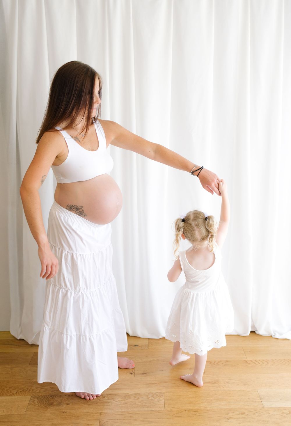 Pregnant woman in white crop top and skirt dancing with toddler girl in matching white dress on hardwood floor.