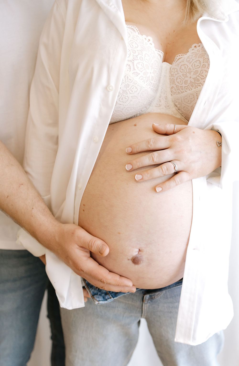 Pregnant woman in white shirt and lace bra with partner's hands cradling baby bump, maternity photo.