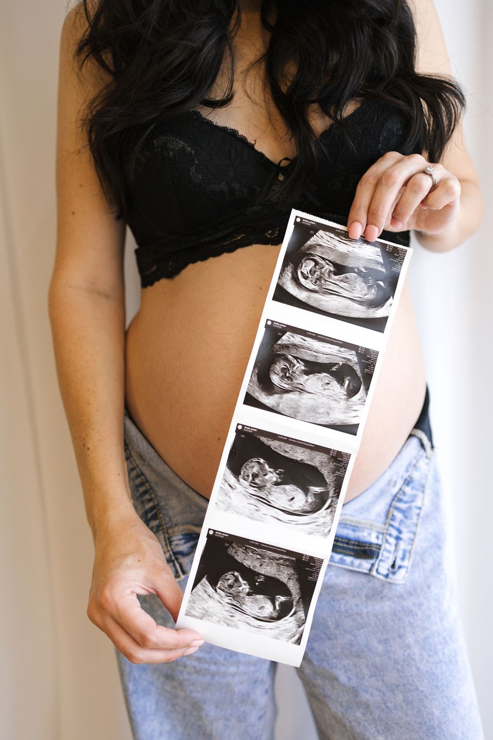 Pregnant woman in black lace bra and jeans holding a strip of four ultrasound sonogram images of her baby.