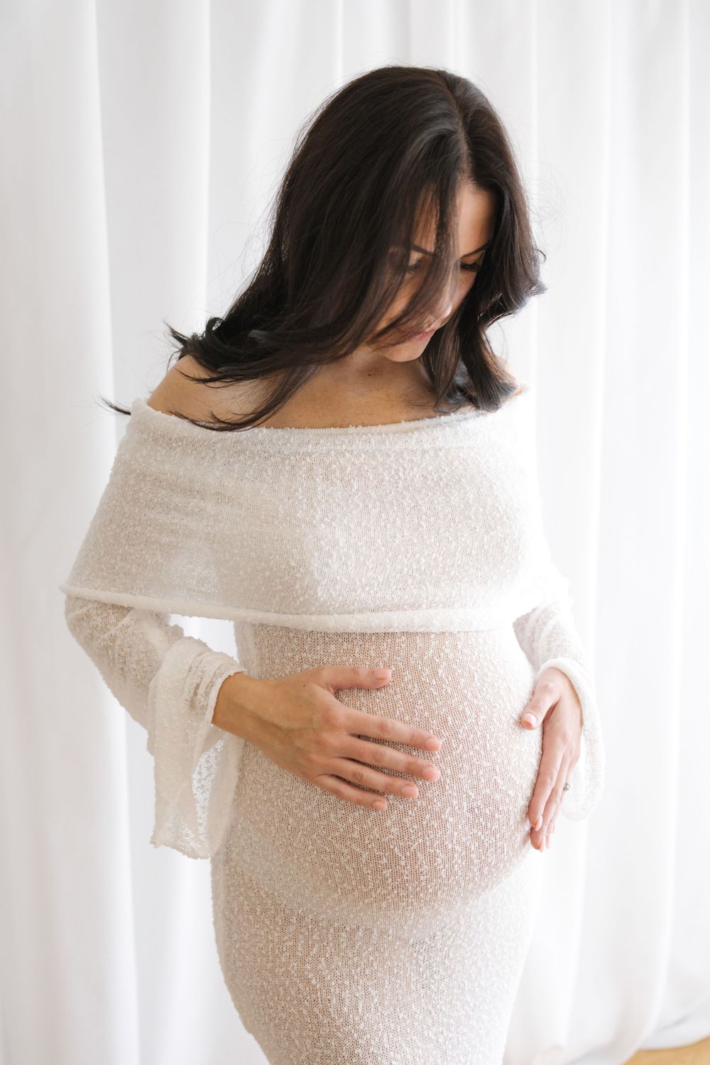 Pregnant woman in white lace off-shoulder dress cradling her baby bump, posing against soft white curtains.