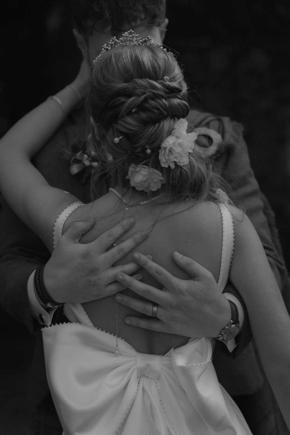Black and white wedding photo of bride with floral updo being embraced by groom, showing elegant backless dress with bow.