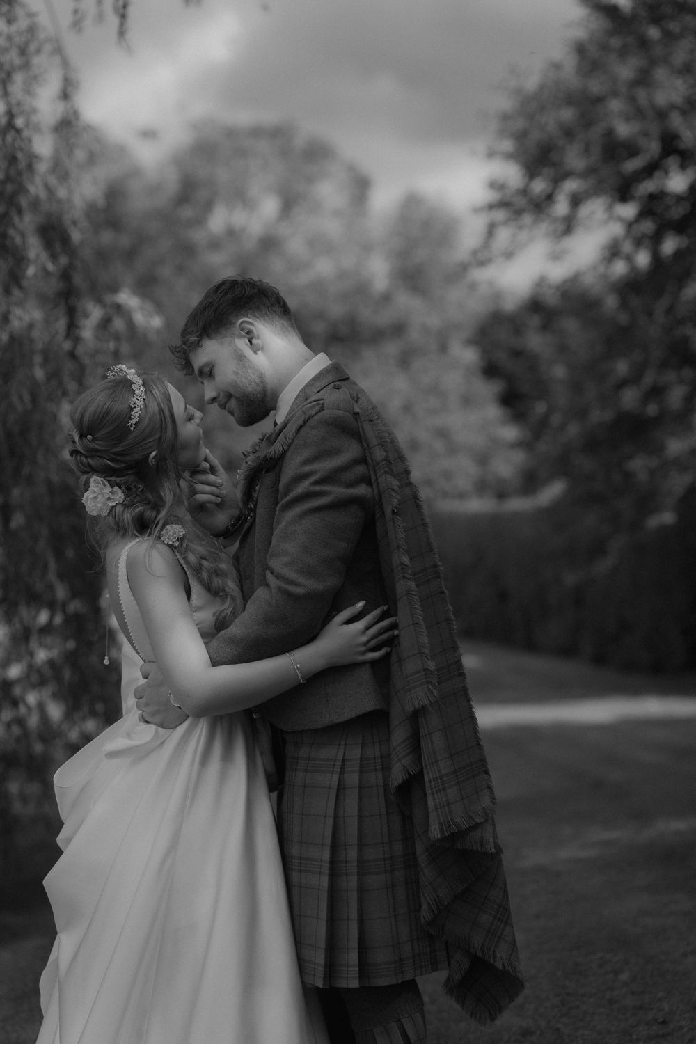 Black and white photo of bride and groom embracing tenderly outdoors, groom wearing traditional Scottish kilt.