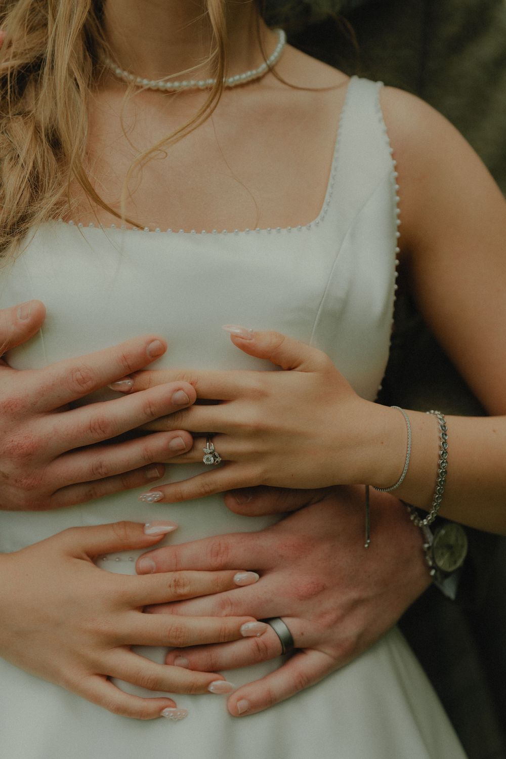 Bride in white dress with pearl necklace and ring, groom embracing her from behind wearing wedding band.
