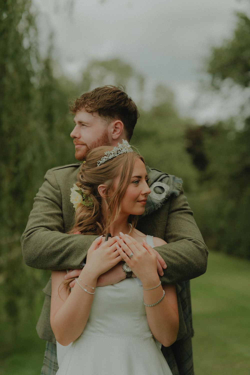 Groom in green tweed jacket embraces bride from behind in lush garden setting on wedding day.