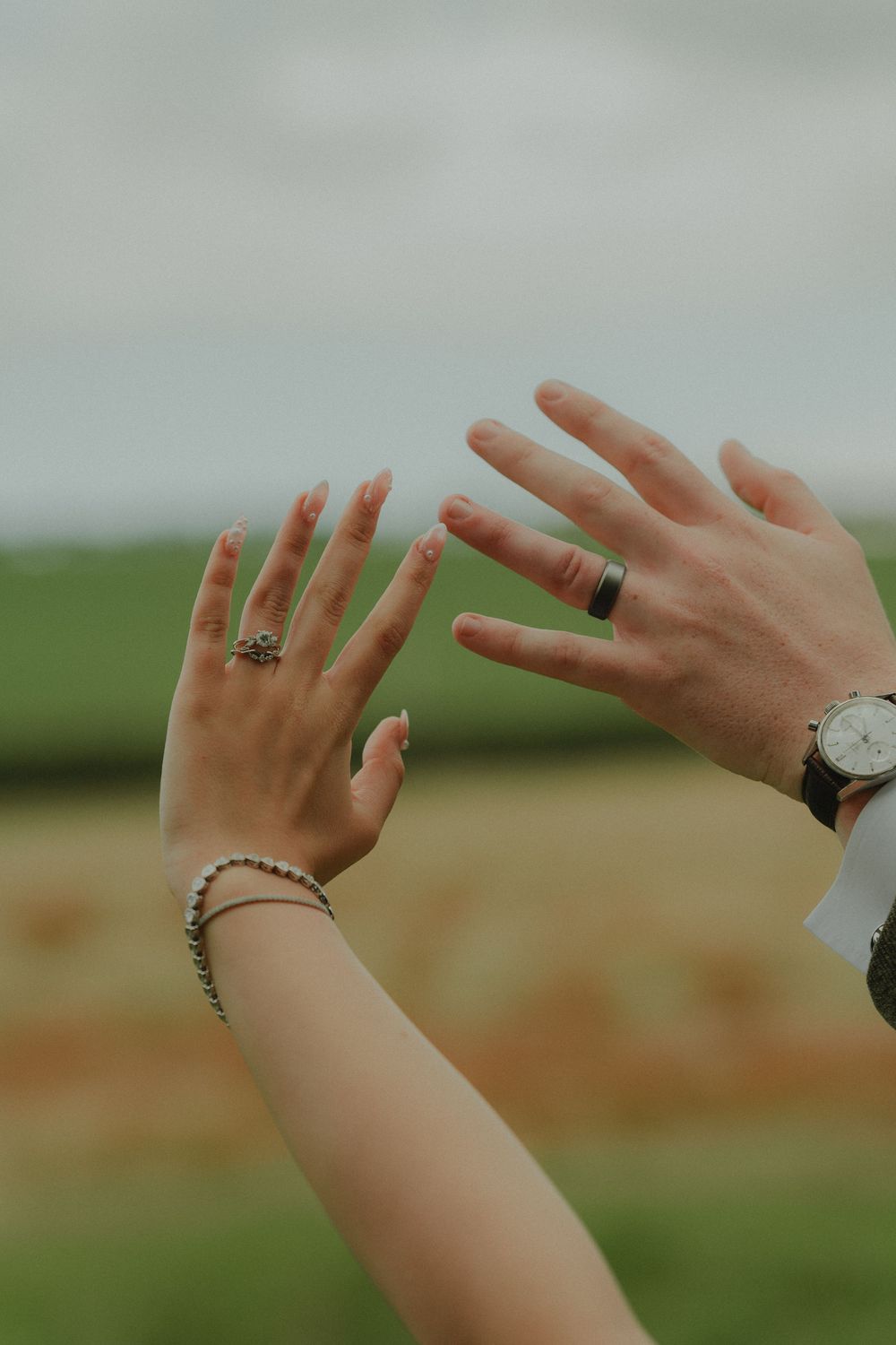 Close-up of bride and groom raising hands showing wedding rings, bracelet, and watch against green field backdrop.