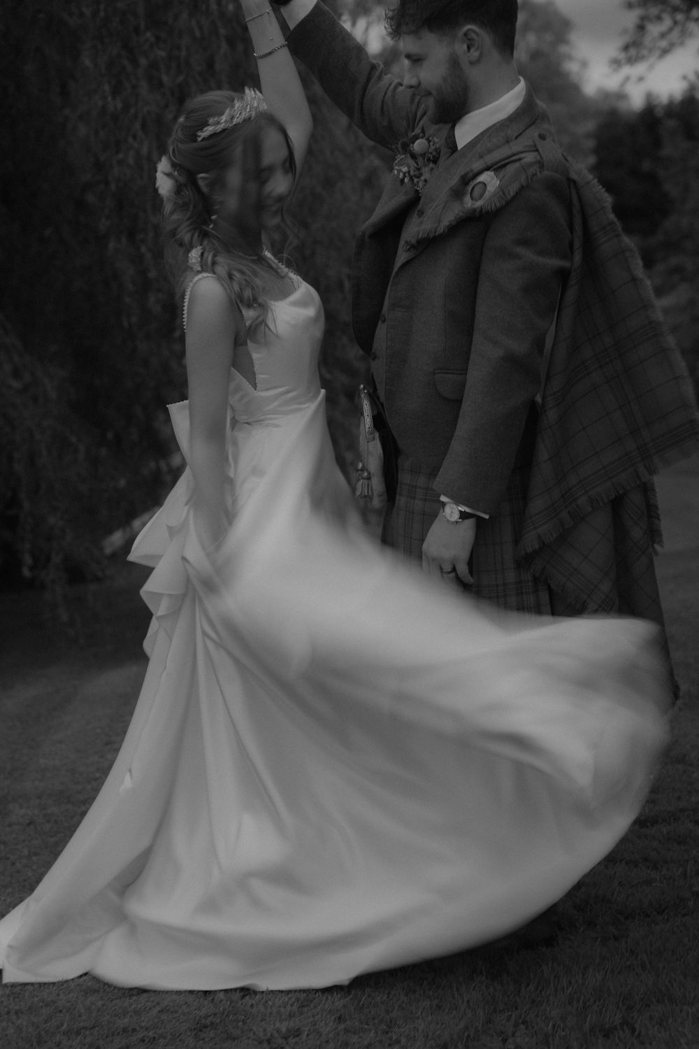 Black and white photo of groom in Scottish tartan kilt twirling bride in flowing white wedding dress outdoors.