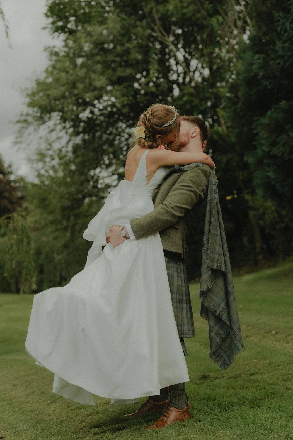 A bride and groom share a romantic kiss outdoors, with the groom wearing a green tartan kilt at their Scottish wedding.