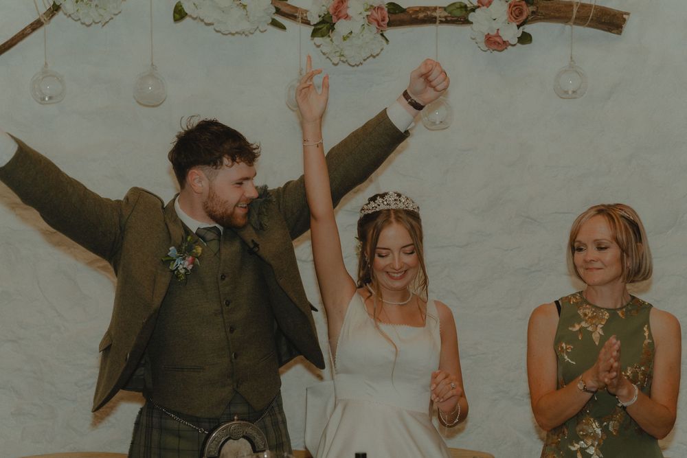 Joyful newlyweds celebrate with raised arms during wedding ceremony beside smiling officiant.