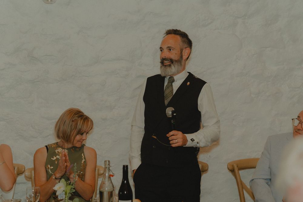 Bearded man in vest giving a speech at a wedding reception, holding a microphone with guests seated nearby.