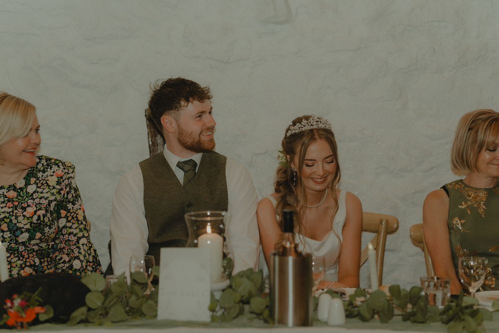 Bride and groom laughing at wedding reception table with guests, floral greenery decor and candles.