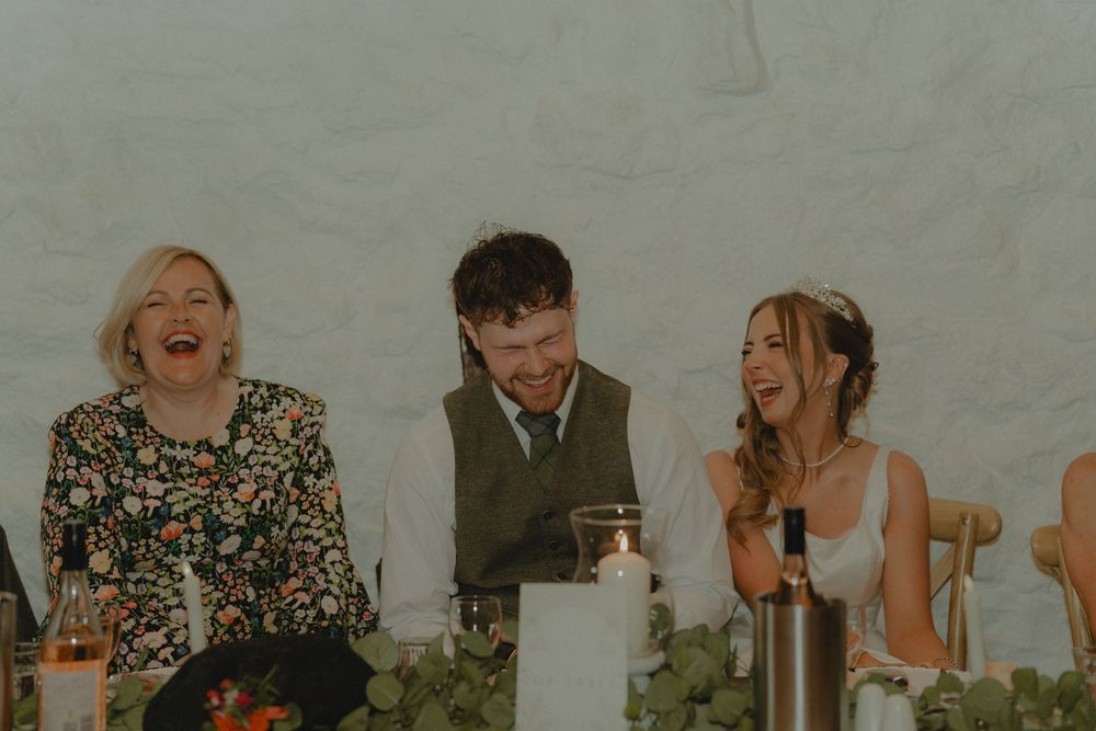 Bride and groom laughing with a guest at the wedding reception dinner table with candles and greenery.